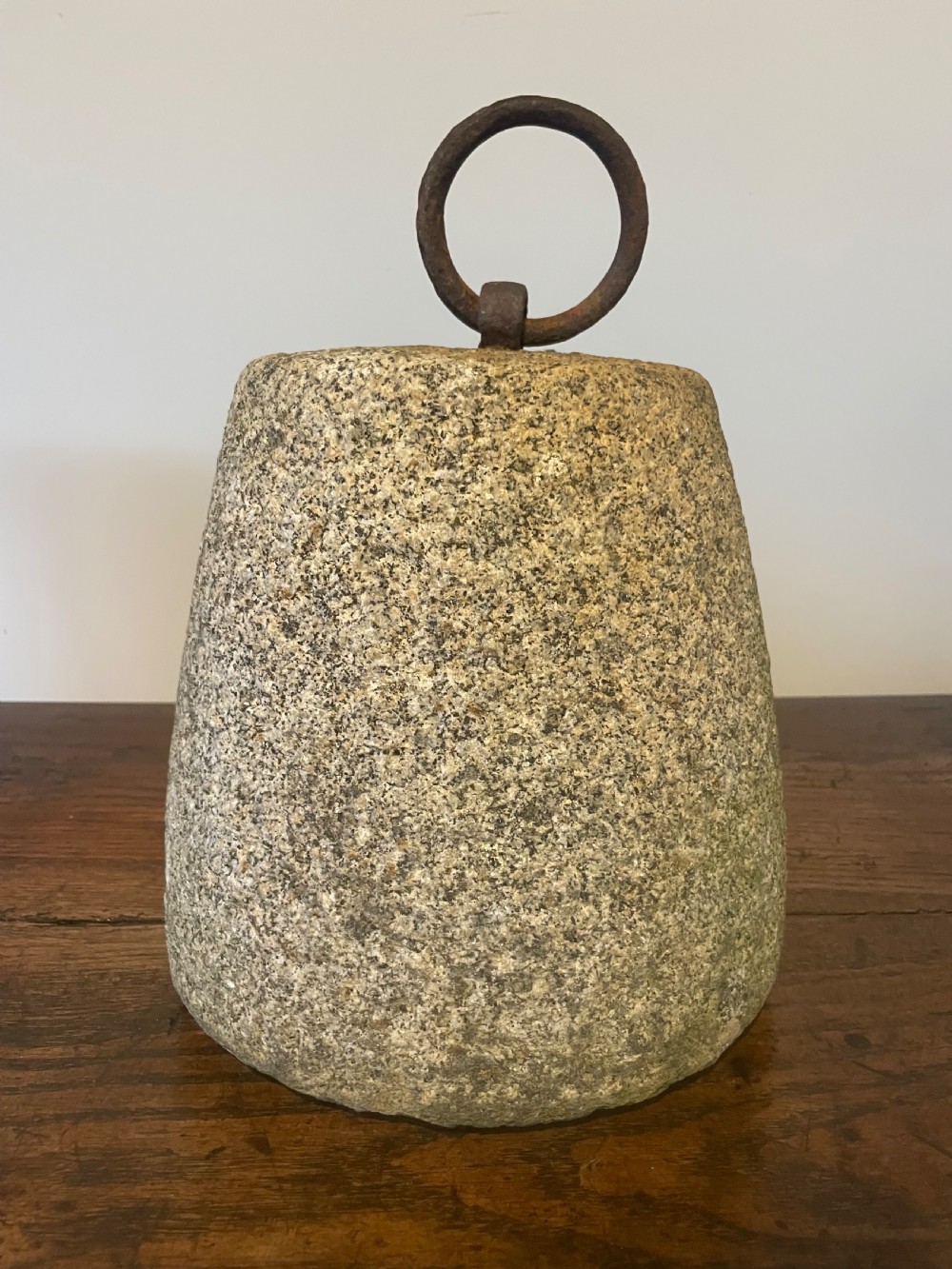 A weathered, tapered granite farm weight with an embedded rusted iron ring on top, resting on a dark wooden surface. View VI