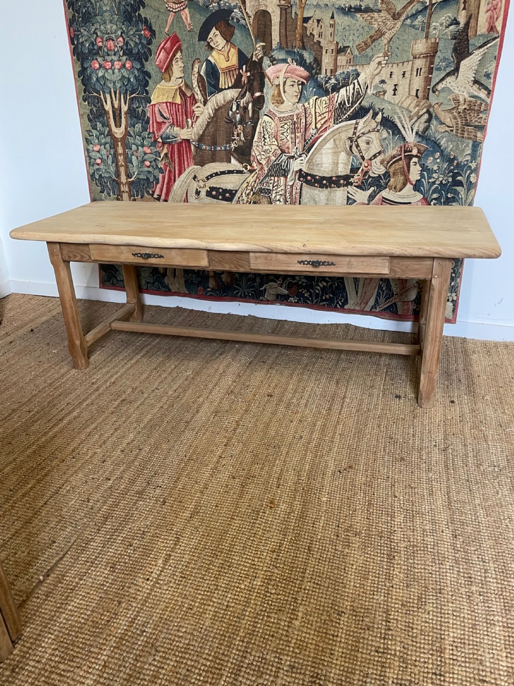 A long light oak farmhouse table featuring a rectangular plank top, two frieze drawers with dark metal handles, and square legs joined by a low stretcher.