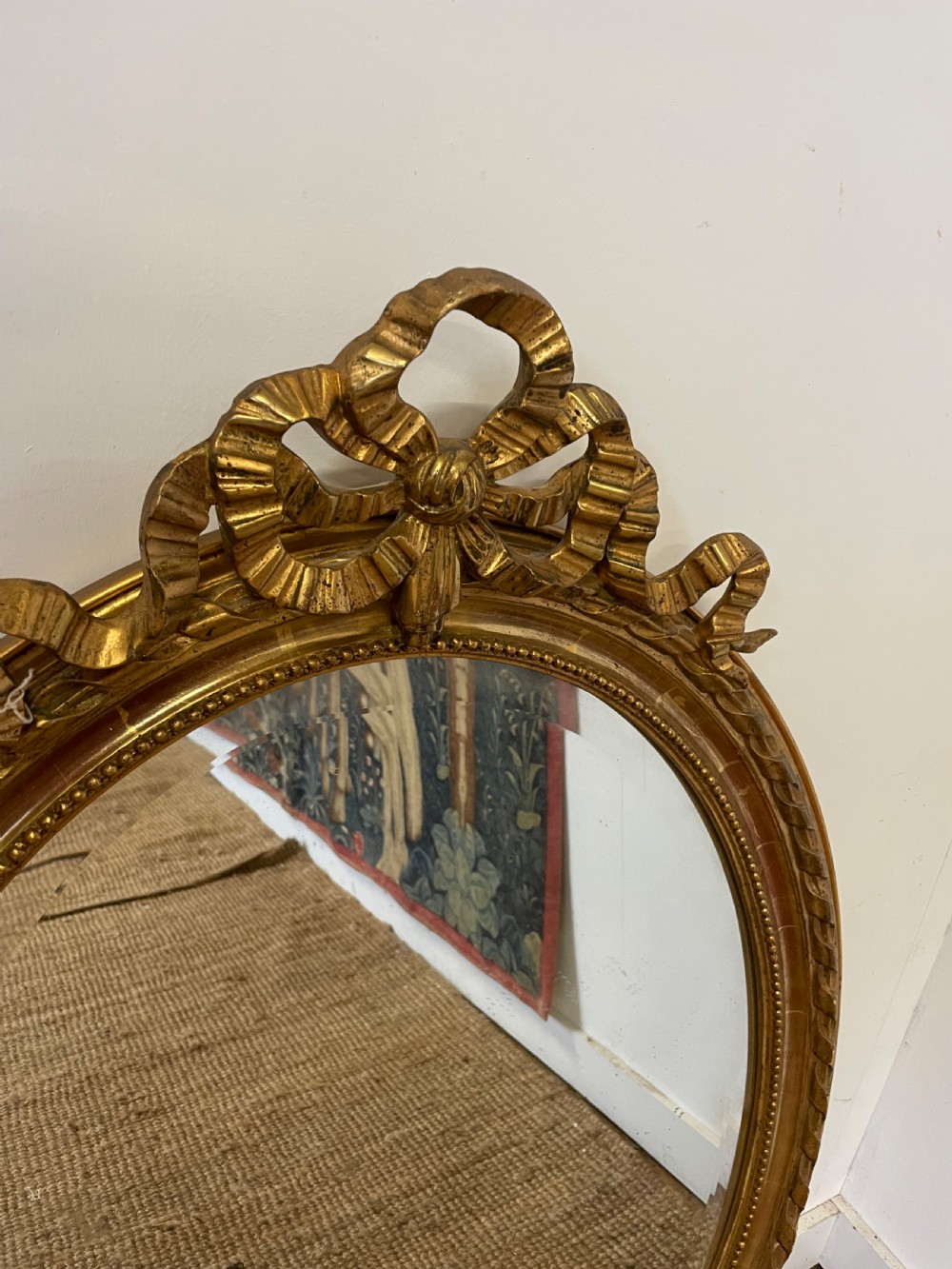 An oval wall mirror featuring a carved gold gilt frame topped with an elaborate ribbon bow scroll. View II
