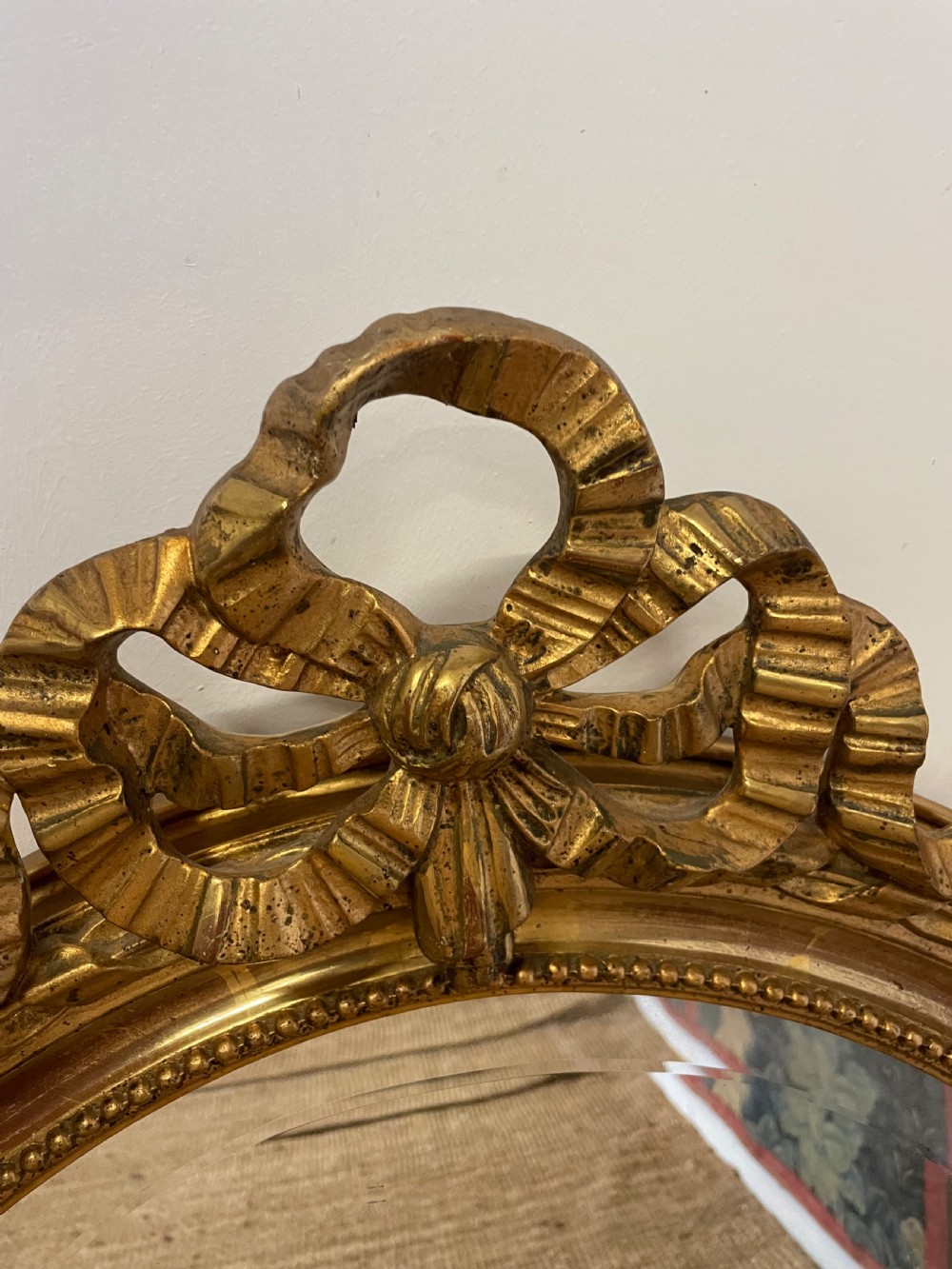 An oval wall mirror featuring a carved gold gilt frame topped with an elaborate ribbon bow scroll. View V