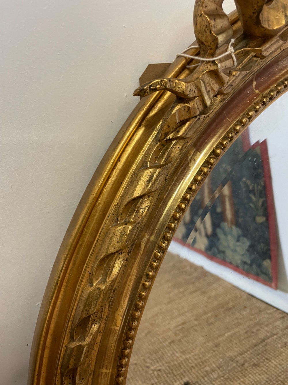 An oval wall mirror featuring a carved gold gilt frame topped with an elaborate ribbon bow scroll. View VIII