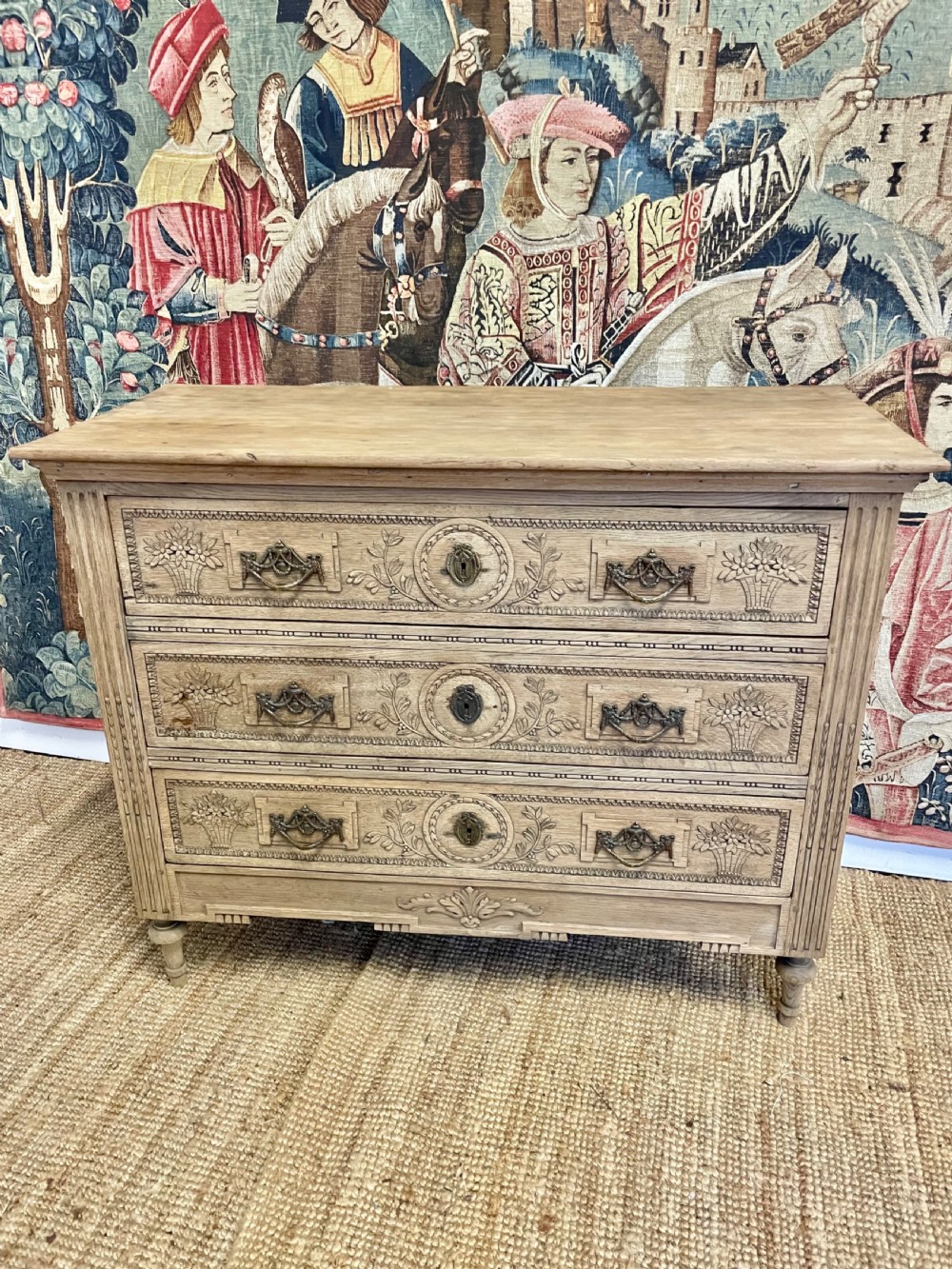 An early 19th-century scrubbed oak commode featuring three hand-carved drawers decorated with floral basket motifs and brass swing handles.