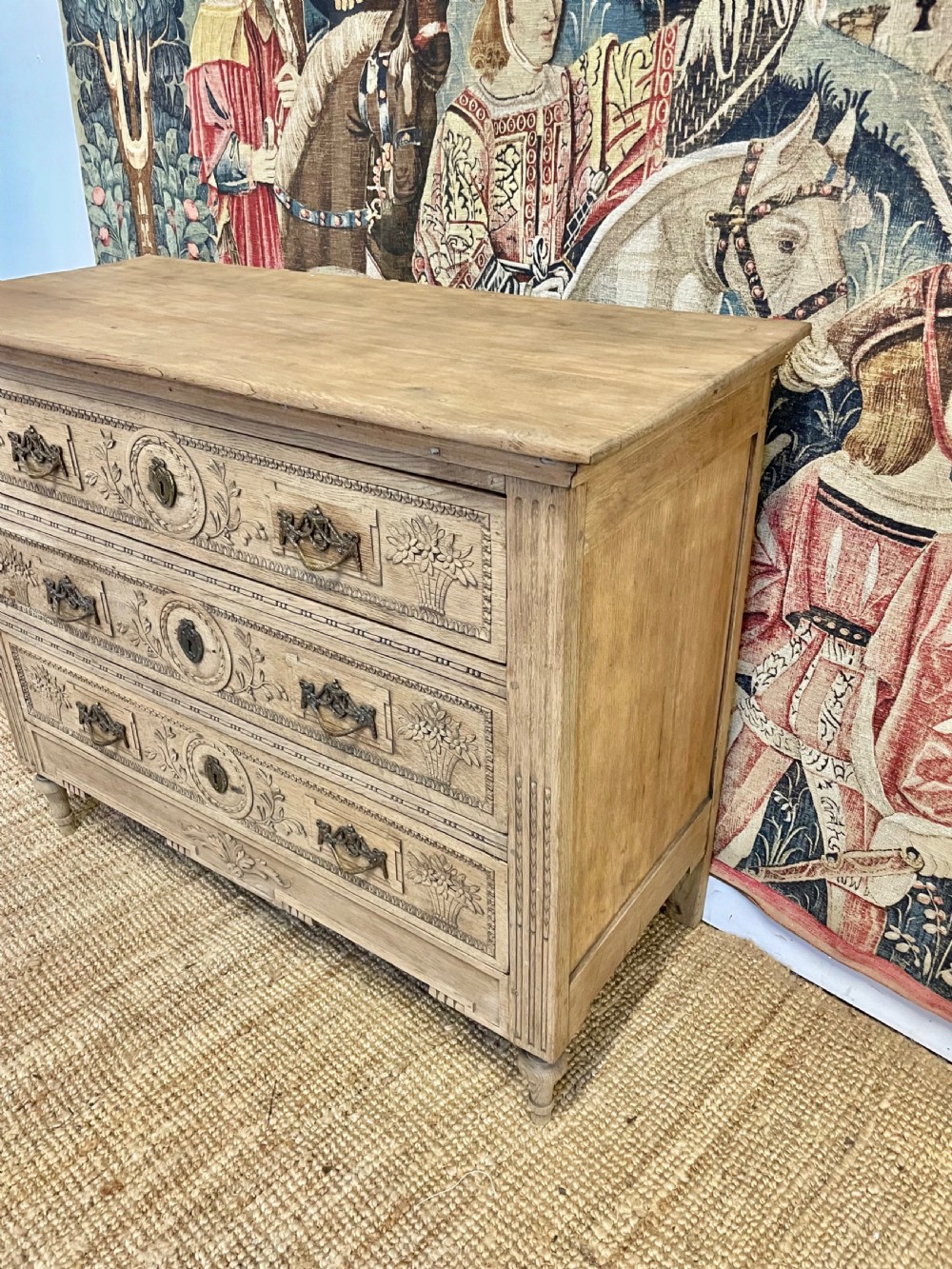 An early 19th-century scrubbed oak commode featuring three hand-carved drawers decorated with floral basket motifs and brass swing handles. View II