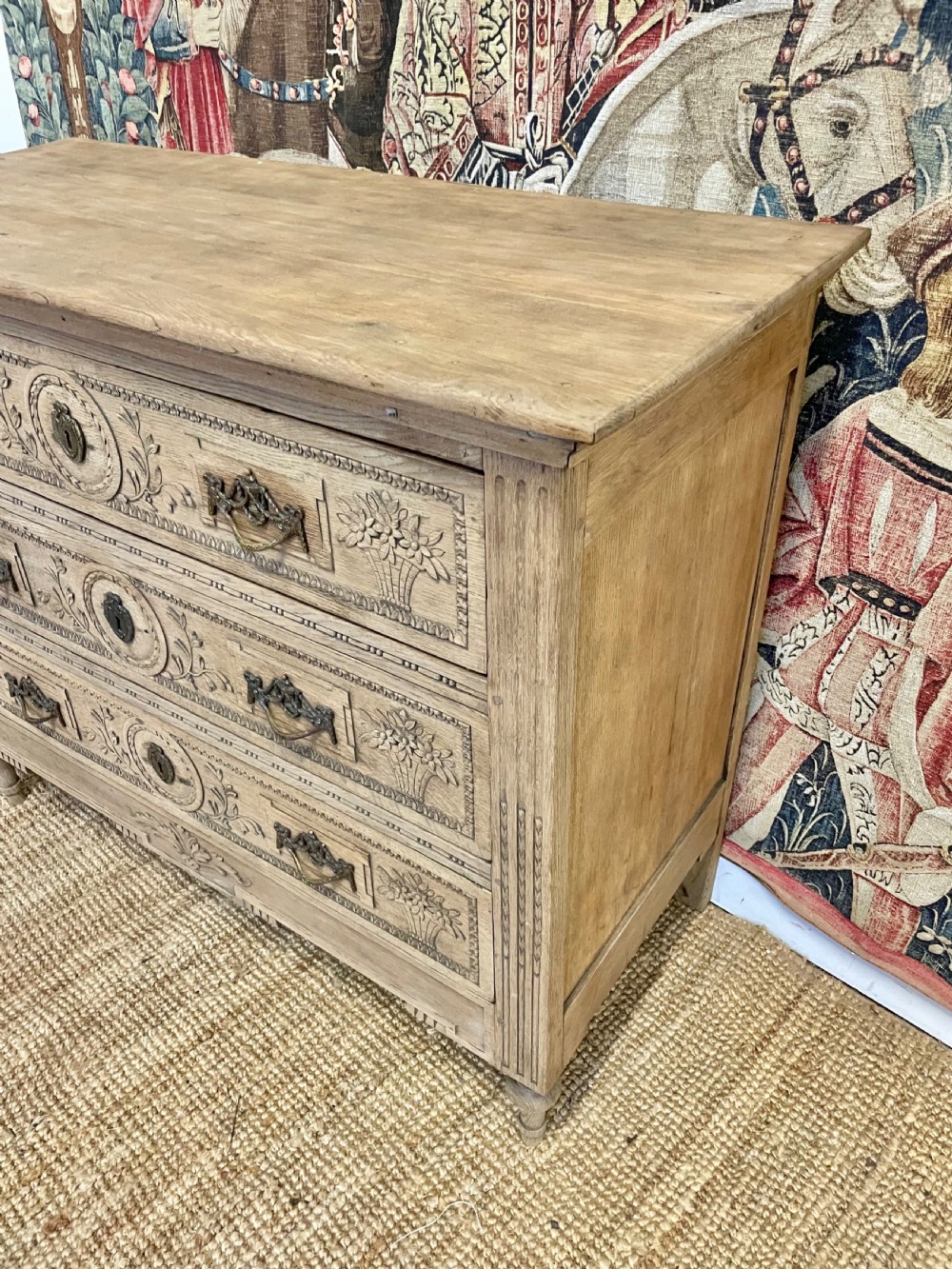 An early 19th-century scrubbed oak commode featuring three hand-carved drawers decorated with floral basket motifs and brass swing handles. View III