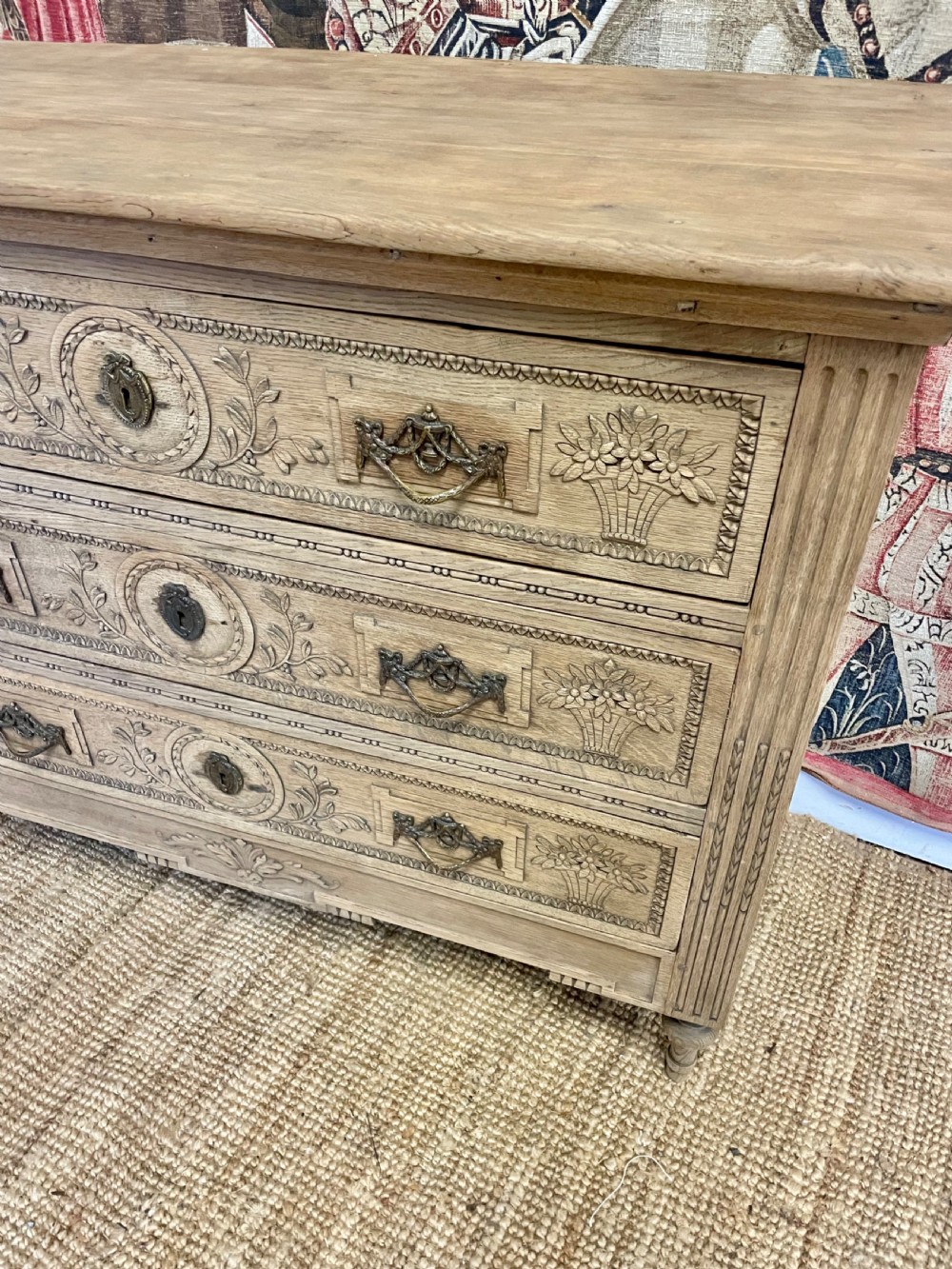 An early 19th-century scrubbed oak commode featuring three hand-carved drawers decorated with floral basket motifs and brass swing handles. View V