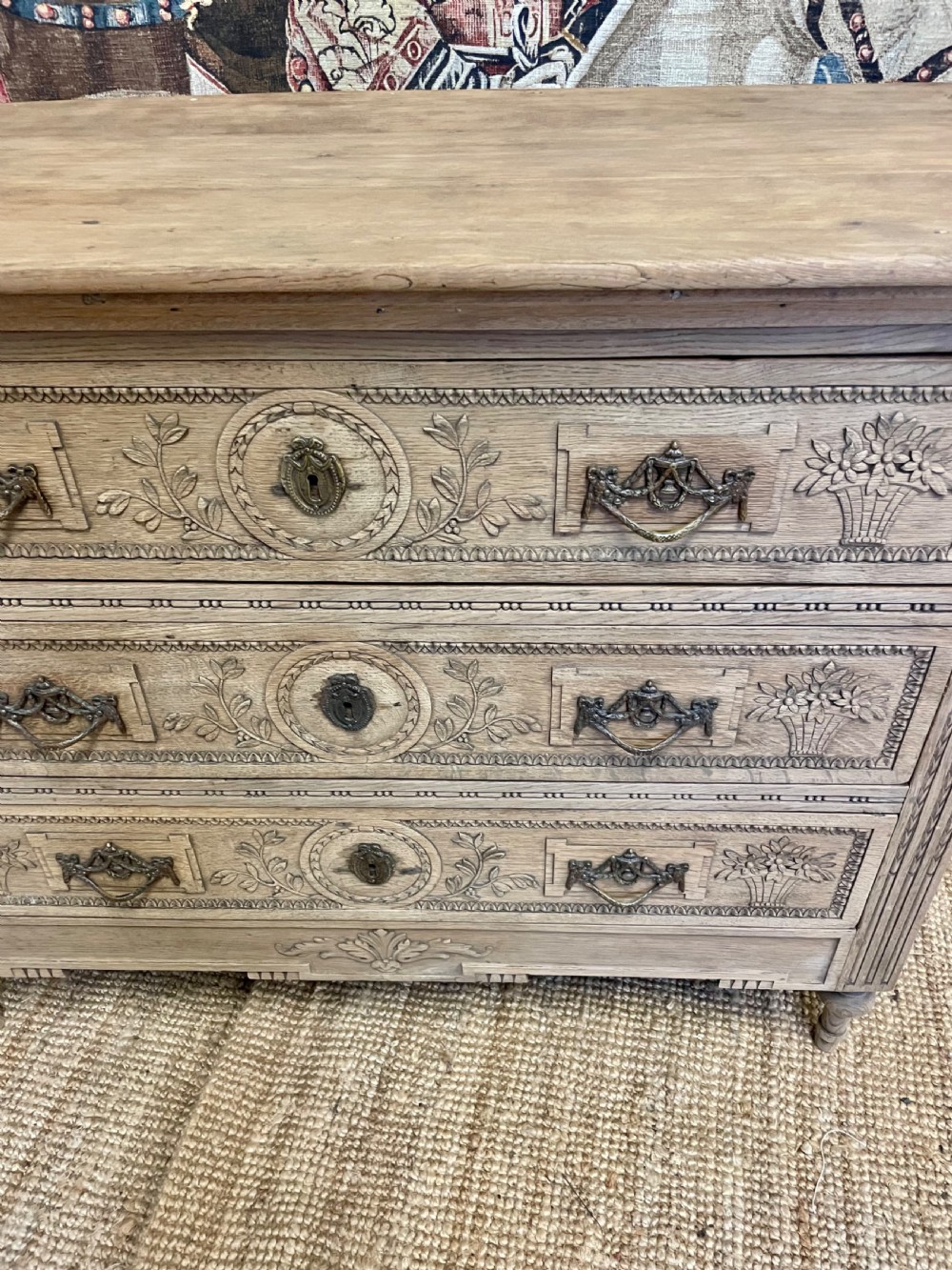 An early 19th-century scrubbed oak commode featuring three hand-carved drawers decorated with floral basket motifs and brass swing handles. View VI