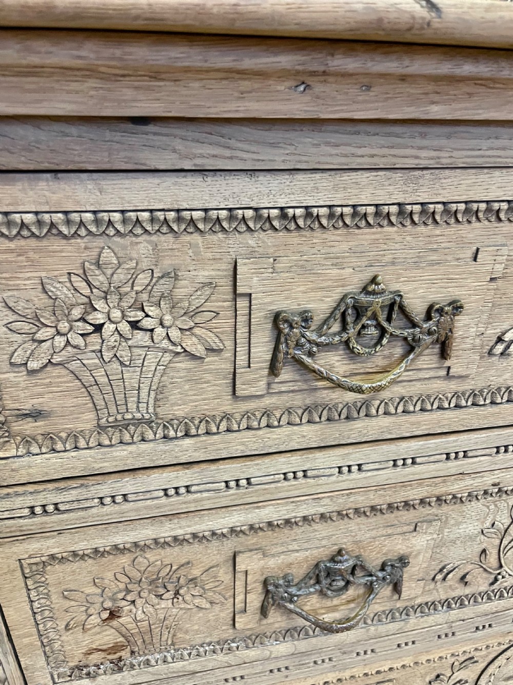 An early 19th-century scrubbed oak commode featuring three hand-carved drawers decorated with floral basket motifs and brass swing handles. View VIII