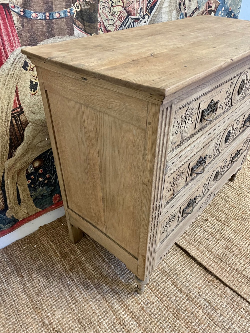An early 19th-century scrubbed oak commode featuring three hand-carved drawers decorated with floral basket motifs and brass swing handles. View X