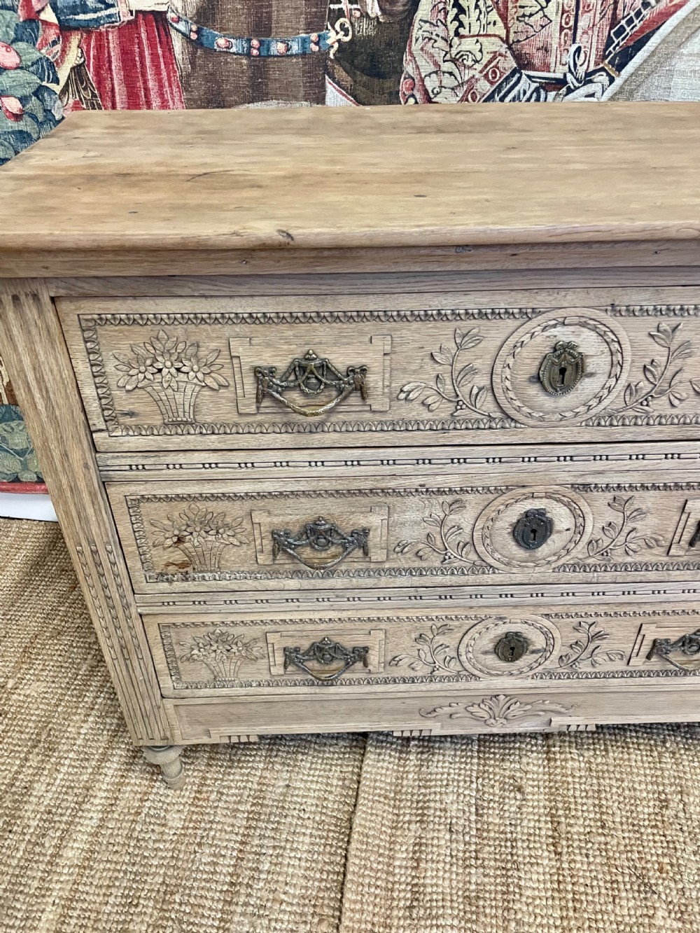An early 19th-century scrubbed oak commode featuring three hand-carved drawers decorated with floral basket motifs and brass swing handles. View XI