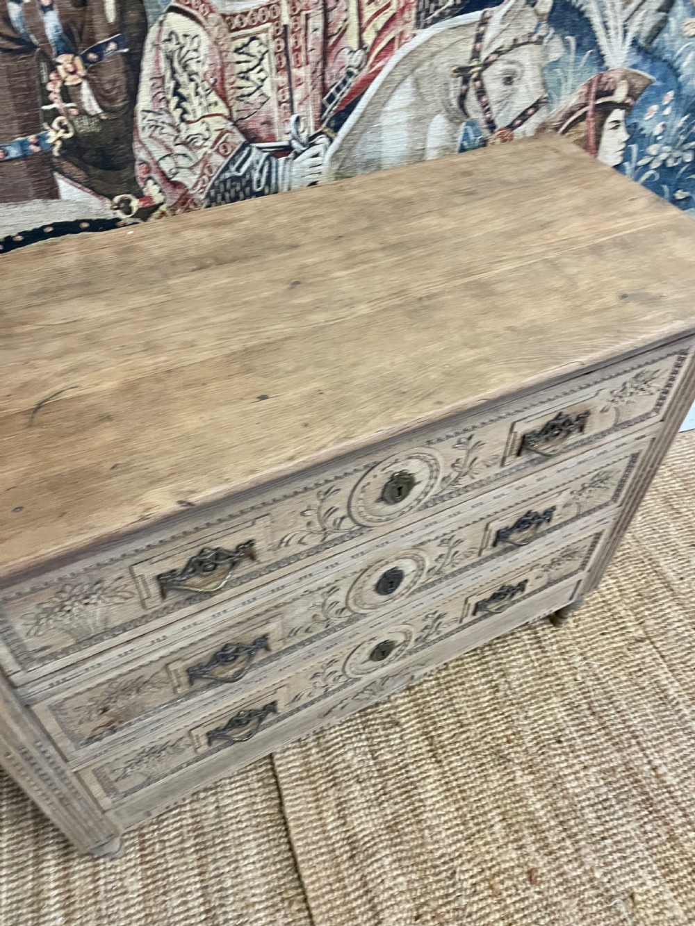 An early 19th-century scrubbed oak commode featuring three hand-carved drawers decorated with floral basket motifs and brass swing handles. View XII
