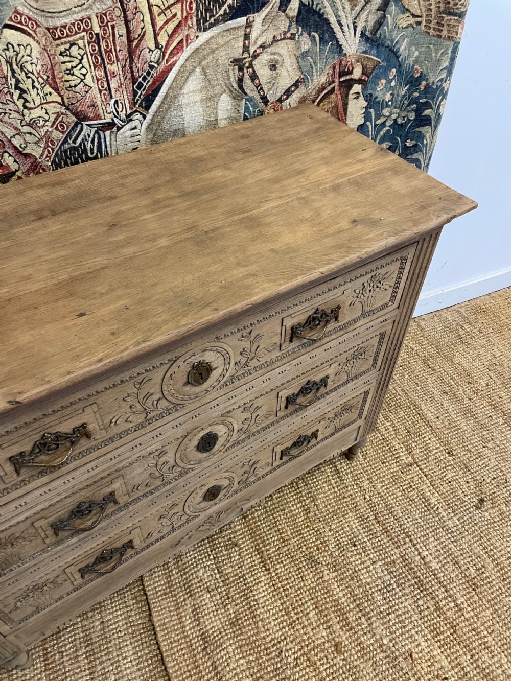 An early 19th-century scrubbed oak commode featuring three hand-carved drawers decorated with floral basket motifs and brass swing handles. View XIII