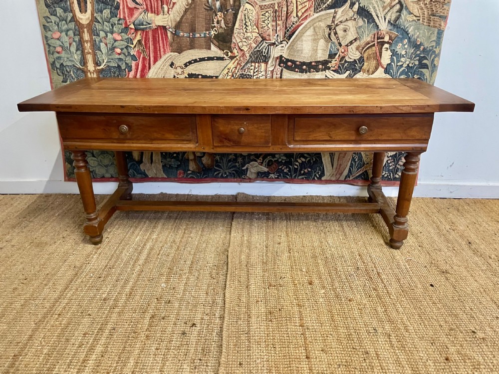 A long rectangular antique cherrywood console table featuring three frieze drawers with small brass knobs and turned legs connected by a low stretcher.