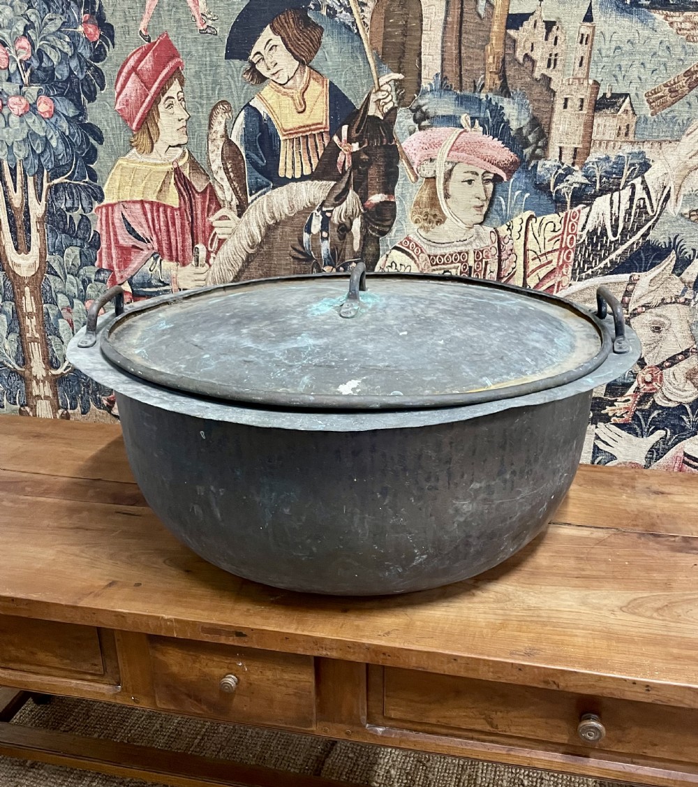 A large round antique copper vessel featuring a fitted lid, twin iron loop handles, and a deep patinated surface with verdigris tones.