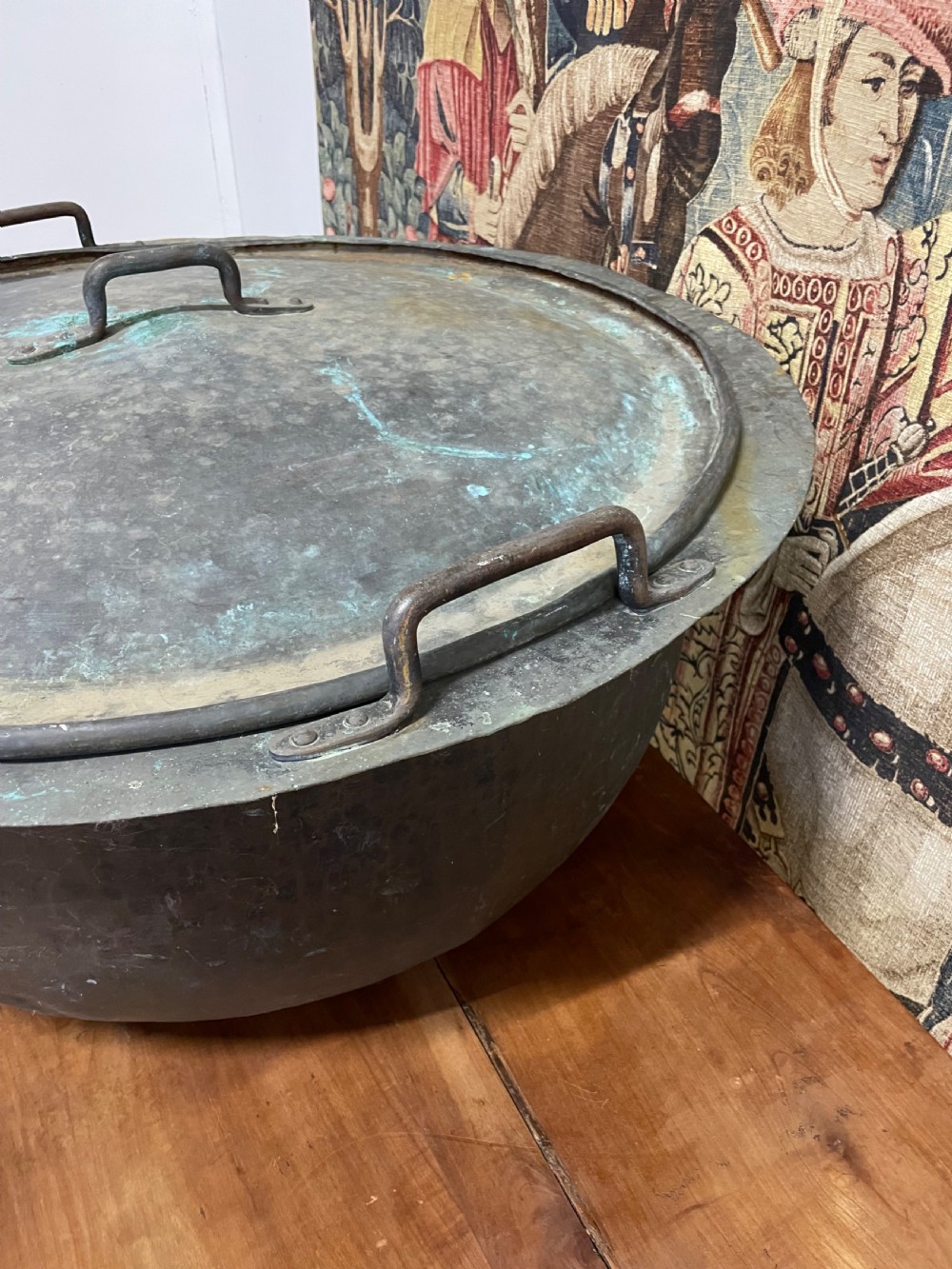 A large round antique copper vessel featuring a fitted lid, twin iron loop handles, and a deep patinated surface with verdigris tones. View V