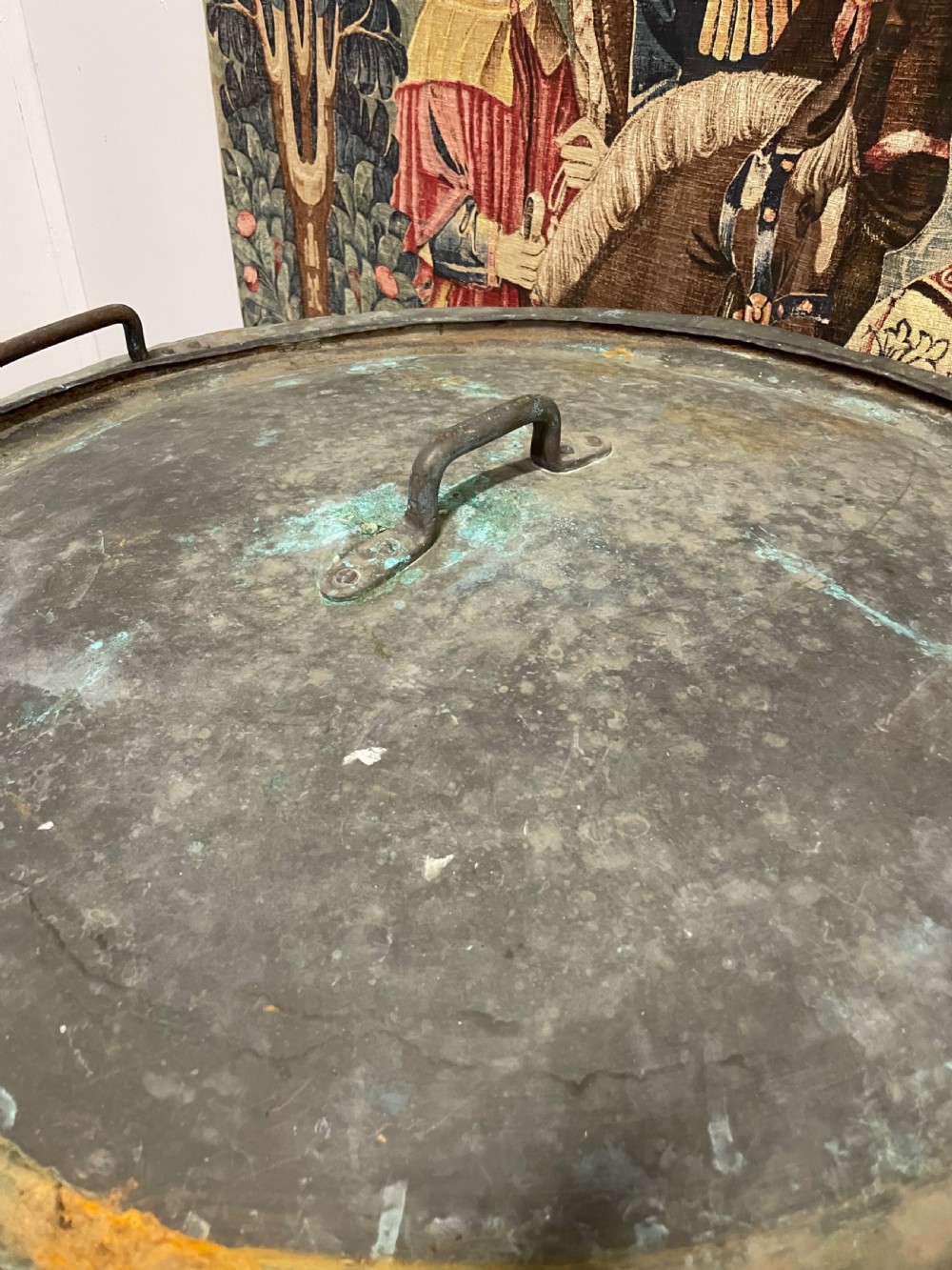 A large round antique copper vessel featuring a fitted lid, twin iron loop handles, and a deep patinated surface with verdigris tones. View VI
