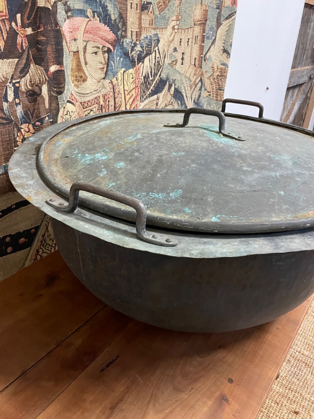 A large round antique copper vessel featuring a fitted lid, twin iron loop handles, and a deep patinated surface with verdigris tones. View VII