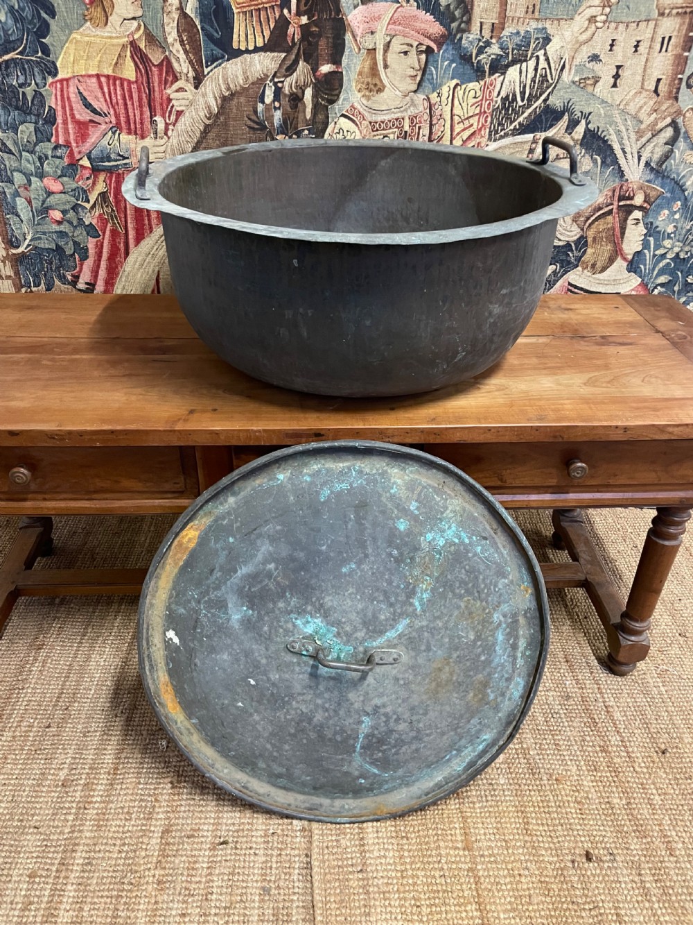 A large round antique copper vessel featuring a fitted lid, twin iron loop handles, and a deep patinated surface with verdigris tones. View IX