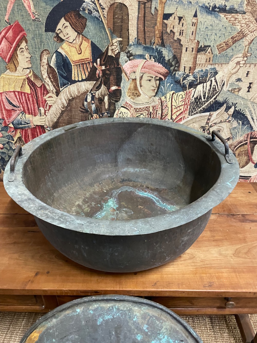 A large round antique copper vessel featuring a fitted lid, twin iron loop handles, and a deep patinated surface with verdigris tones. View X