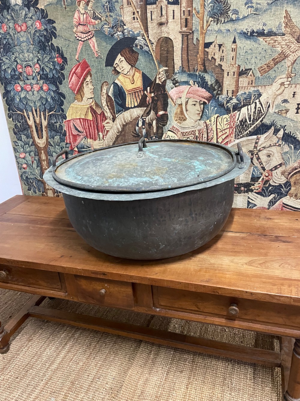 A large round antique copper vessel featuring a fitted lid, twin iron loop handles, and a deep patinated surface with verdigris tones. View XI