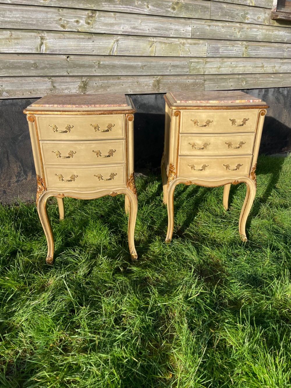 A pair of early 20th century French style cream painted and gilded bedside cabinets featuring fitted marble tops, three drawers, and carved cabriole legs.