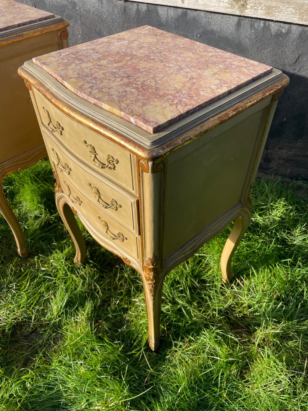 A pair of early 20th century French style cream painted and gilded bedside cabinets featuring fitted marble tops, three drawers, and carved cabriole legs. View V