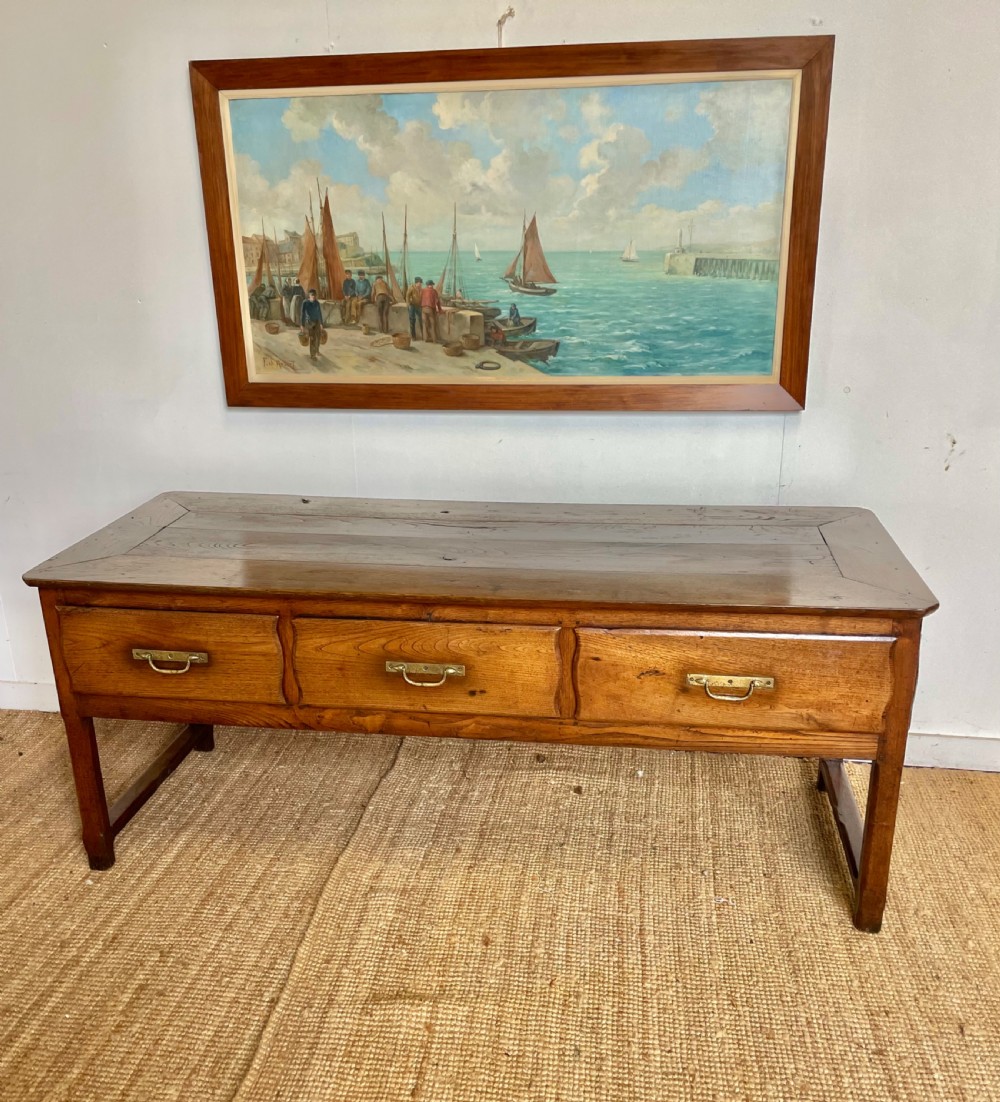 An antique chestnut dresser base features a rectangular wooden top, three horizontal drawers with brass drop handles, and straight legs connected by stretchers.