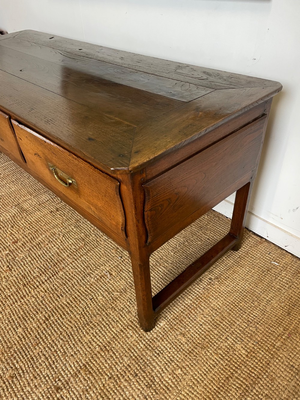 An antique chestnut dresser base features a rectangular wooden top, three horizontal drawers with brass drop handles, and straight legs connected by stretchers. View III