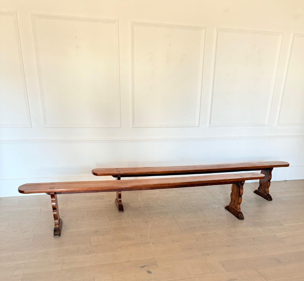 A pair of long 19th-century cherrywood benches with single-plank slab seats and shaped trestle end supports.