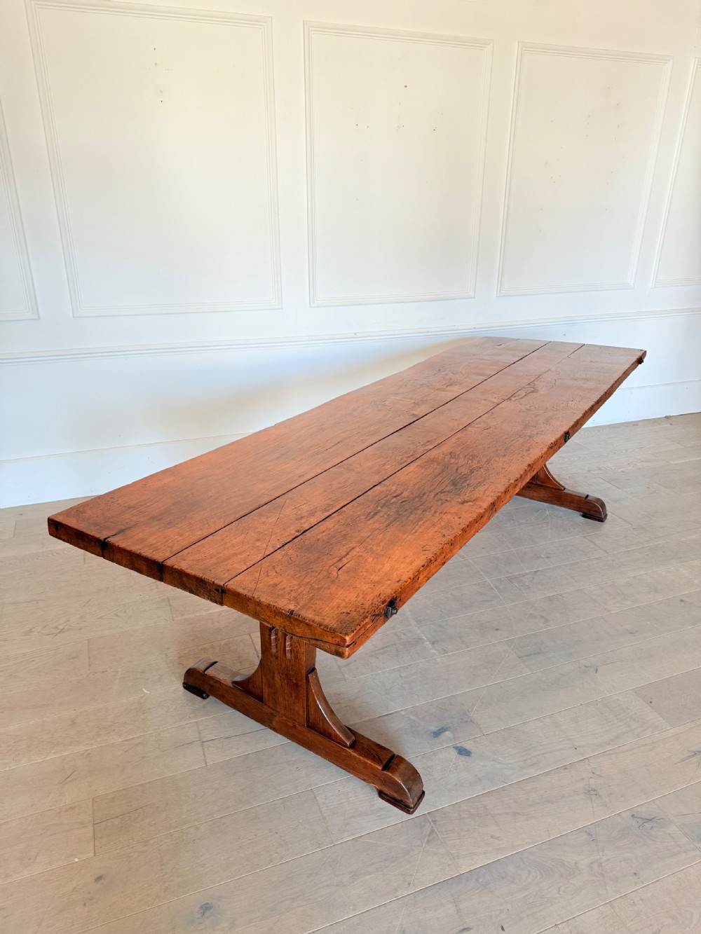 An early 19th century fruitwood refectory table featuring a plank top and trestle base against a white panelled wall.