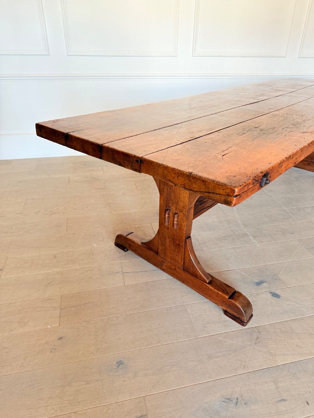An early 19th century fruitwood refectory table featuring a plank top and trestle base against a white panelled wall. View II