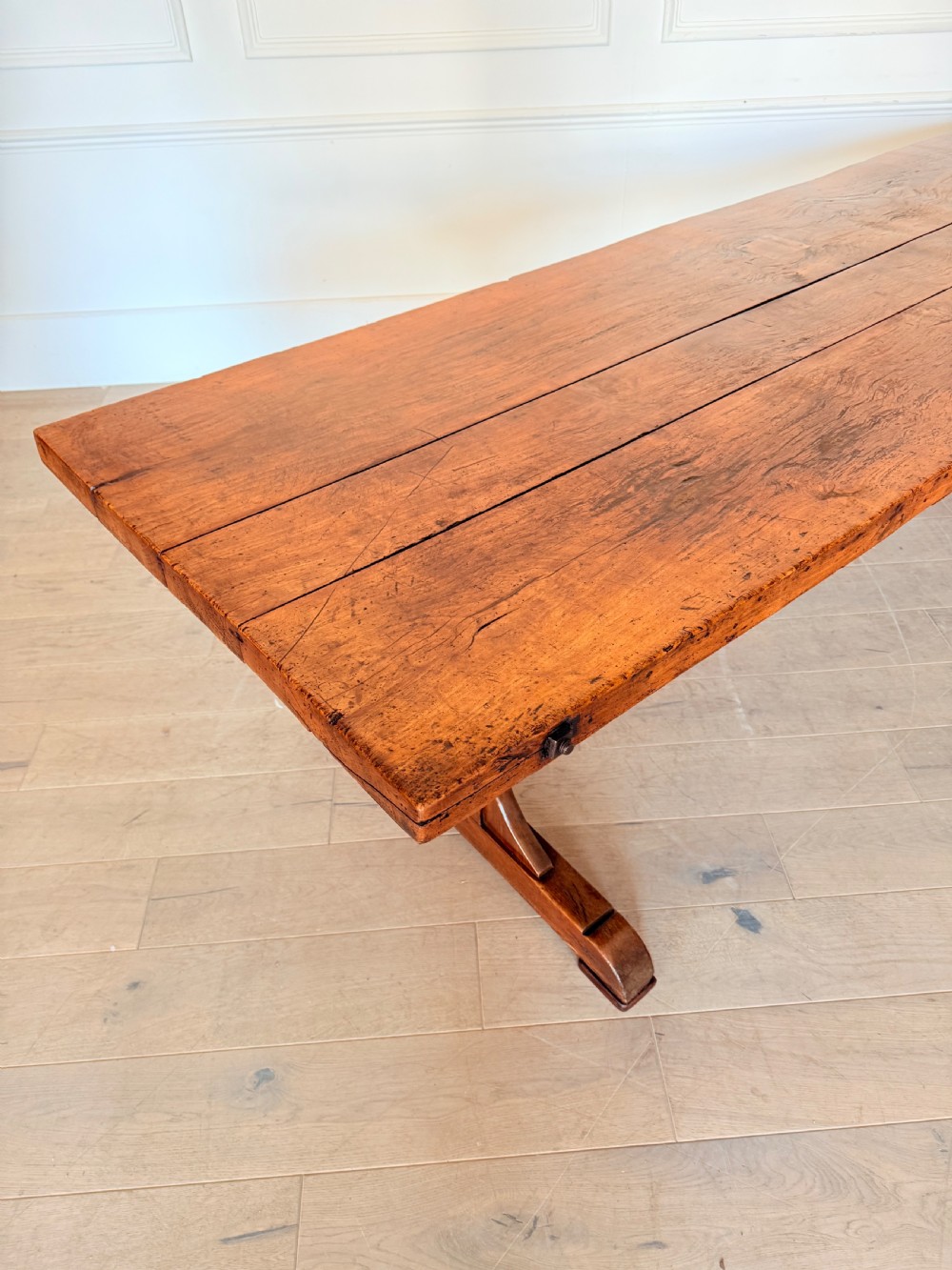 An early 19th century fruitwood refectory table featuring a plank top and trestle base against a white panelled wall. View III