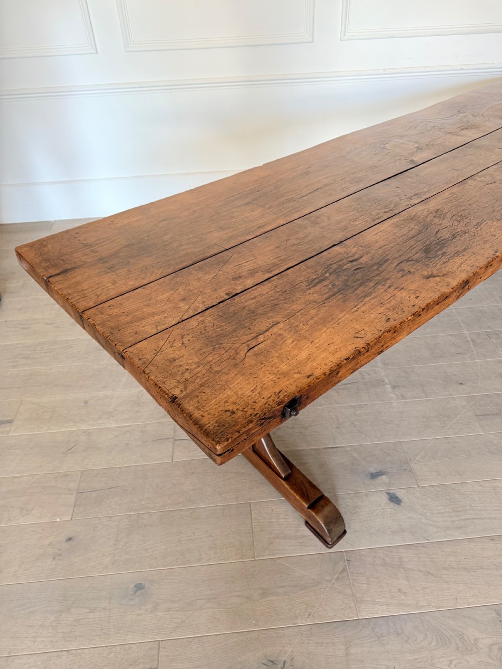 An early 19th century fruitwood refectory table featuring a plank top and trestle base against a white panelled wall. View IV