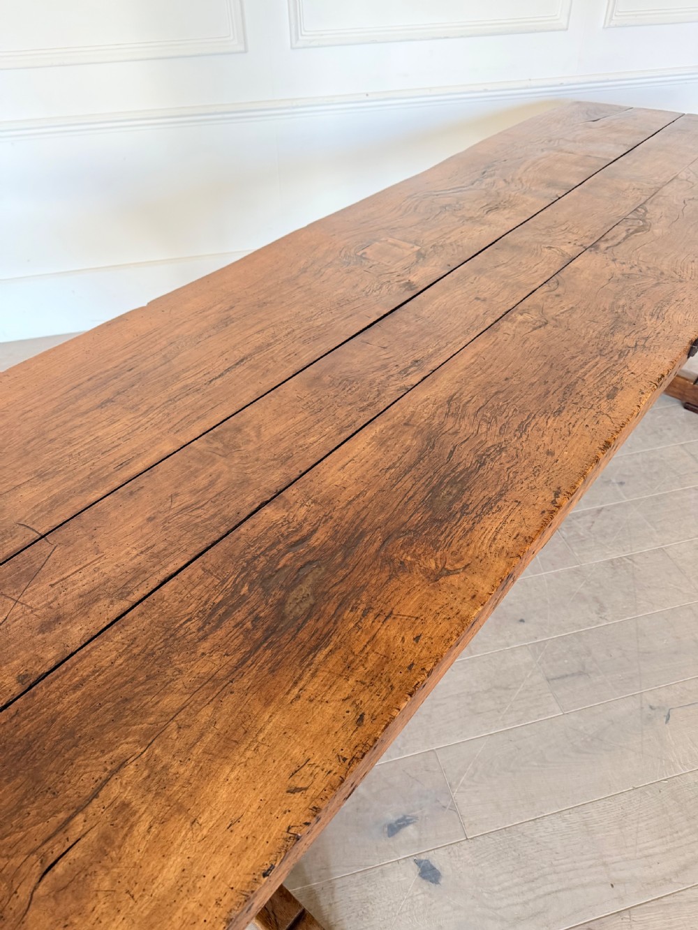 An early 19th century fruitwood refectory table featuring a plank top and trestle base against a white panelled wall. View V