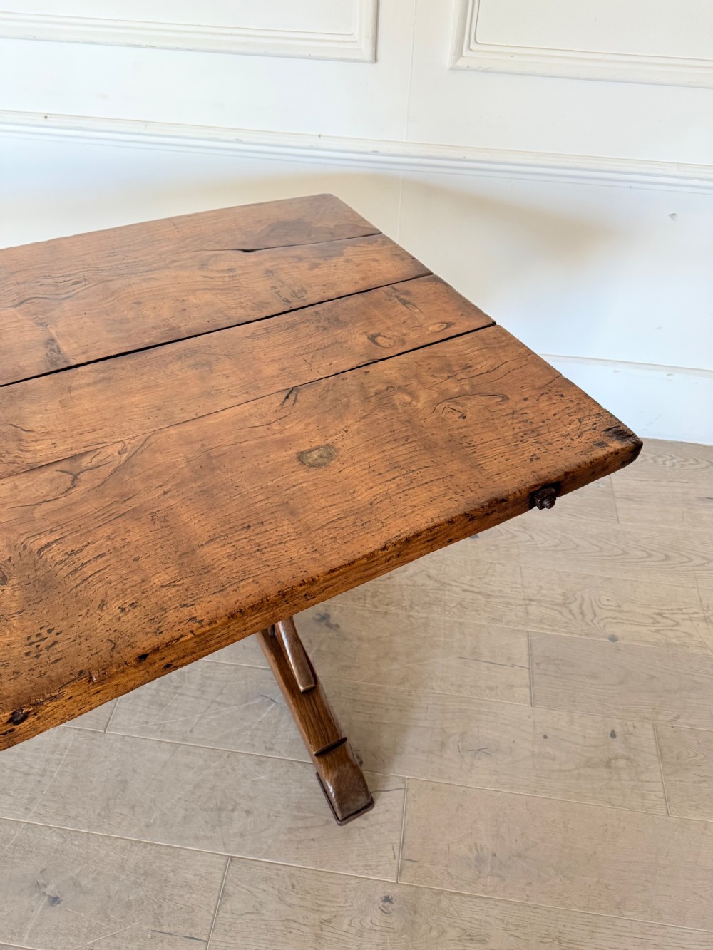 An early 19th century fruitwood refectory table featuring a plank top and trestle base against a white panelled wall. View VIII
