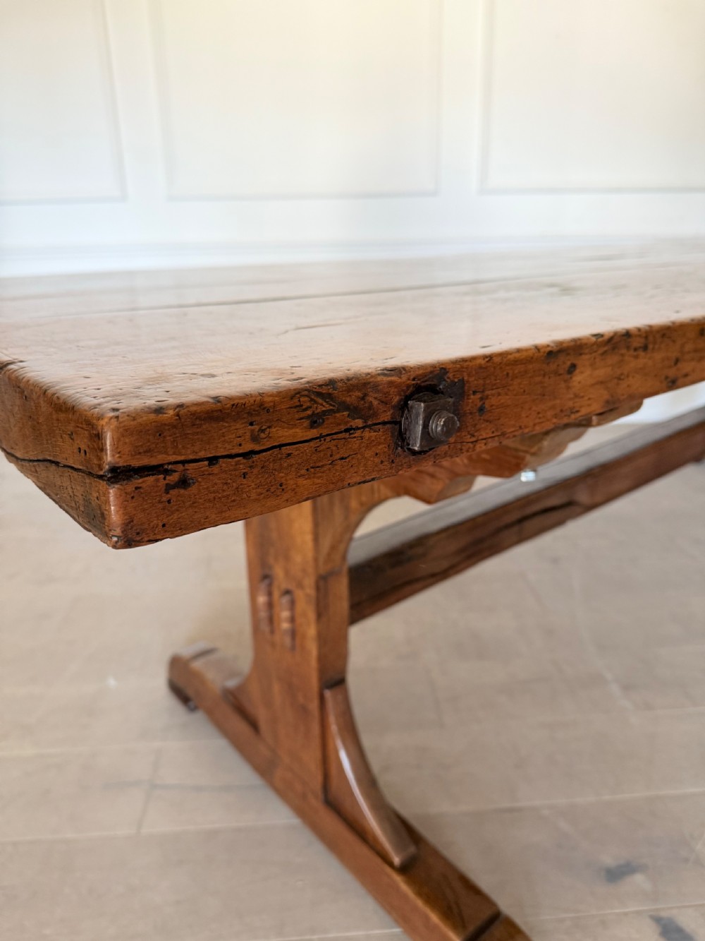 An early 19th century fruitwood refectory table featuring a plank top and trestle base against a white panelled wall. View XVII