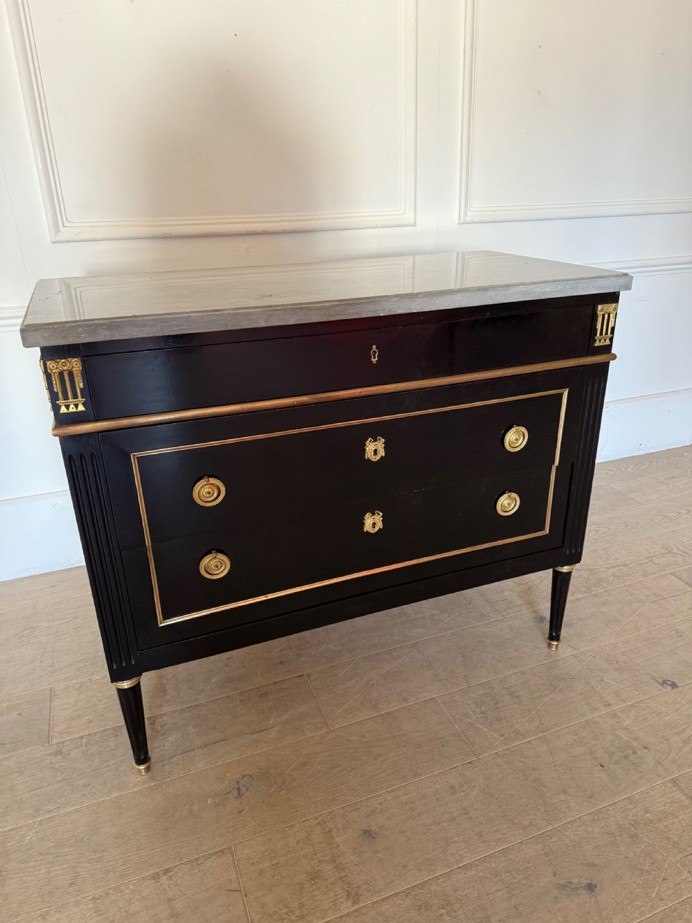 An ebonised commode chest of drawers featuring a grey marble top, brass ring handles, and gilt metal mounts.