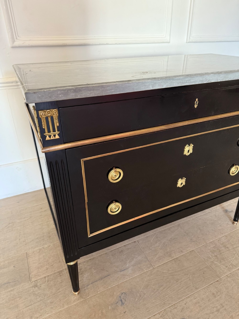 An ebonised commode chest of drawers featuring a grey marble top, brass ring handles, and gilt metal mounts. View II
