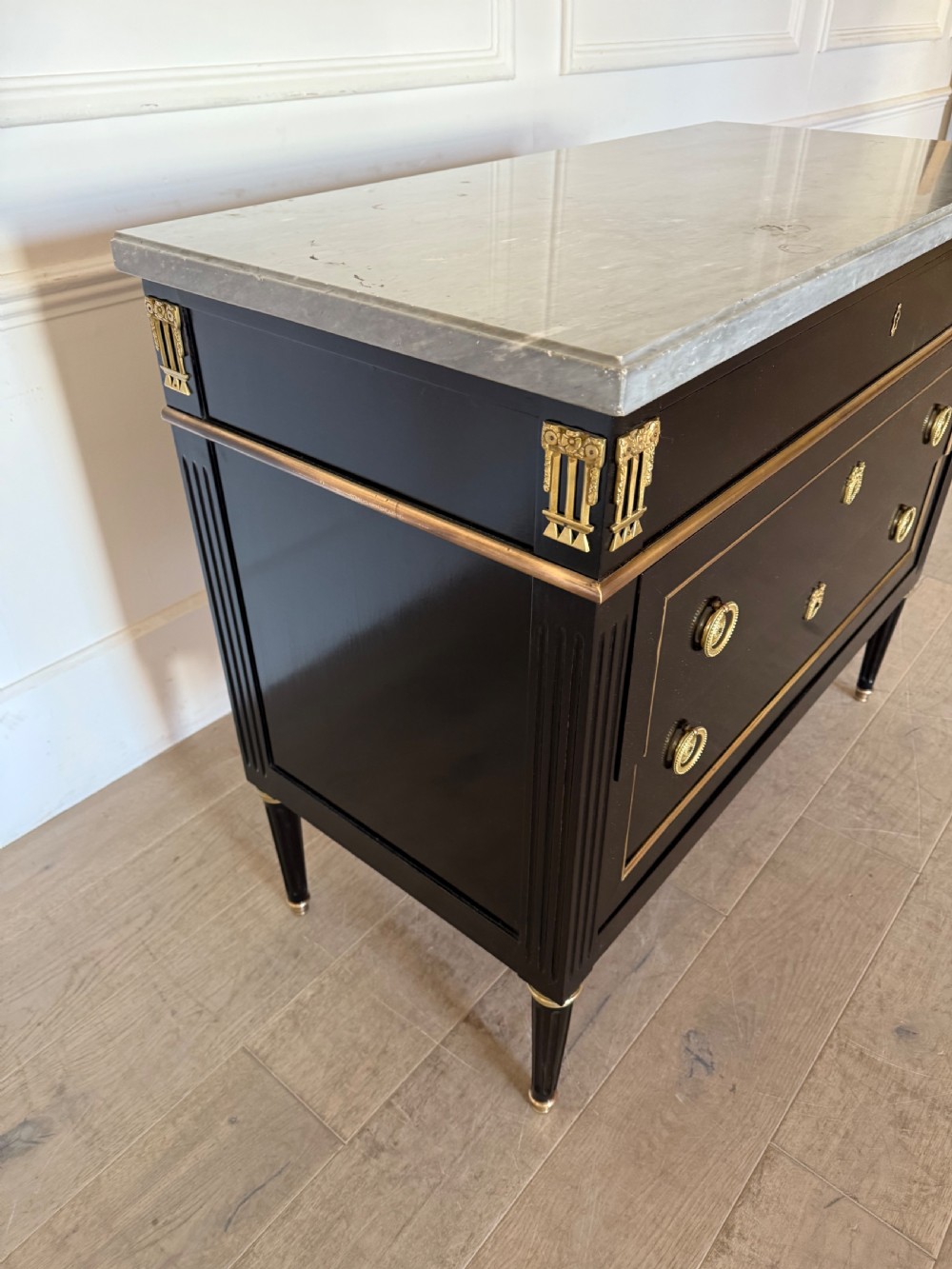An ebonised commode chest of drawers featuring a grey marble top, brass ring handles, and gilt metal mounts. View III