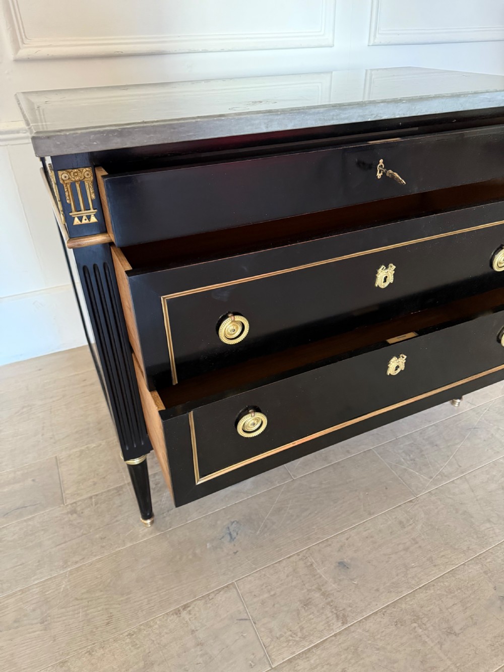 An ebonised commode chest of drawers featuring a grey marble top, brass ring handles, and gilt metal mounts. View IV