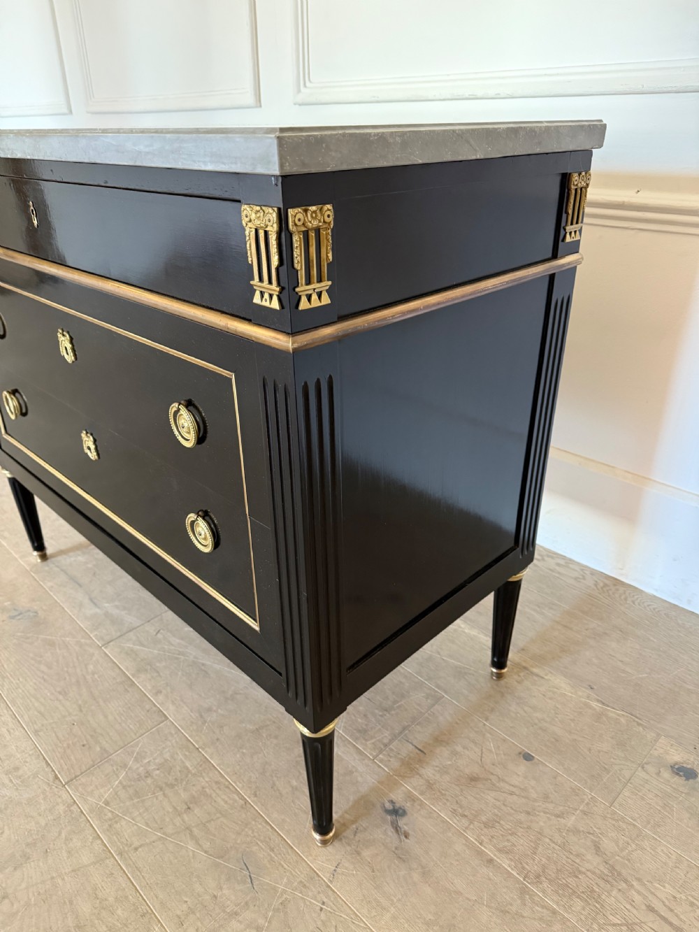 An ebonised commode chest of drawers featuring a grey marble top, brass ring handles, and gilt metal mounts. View V