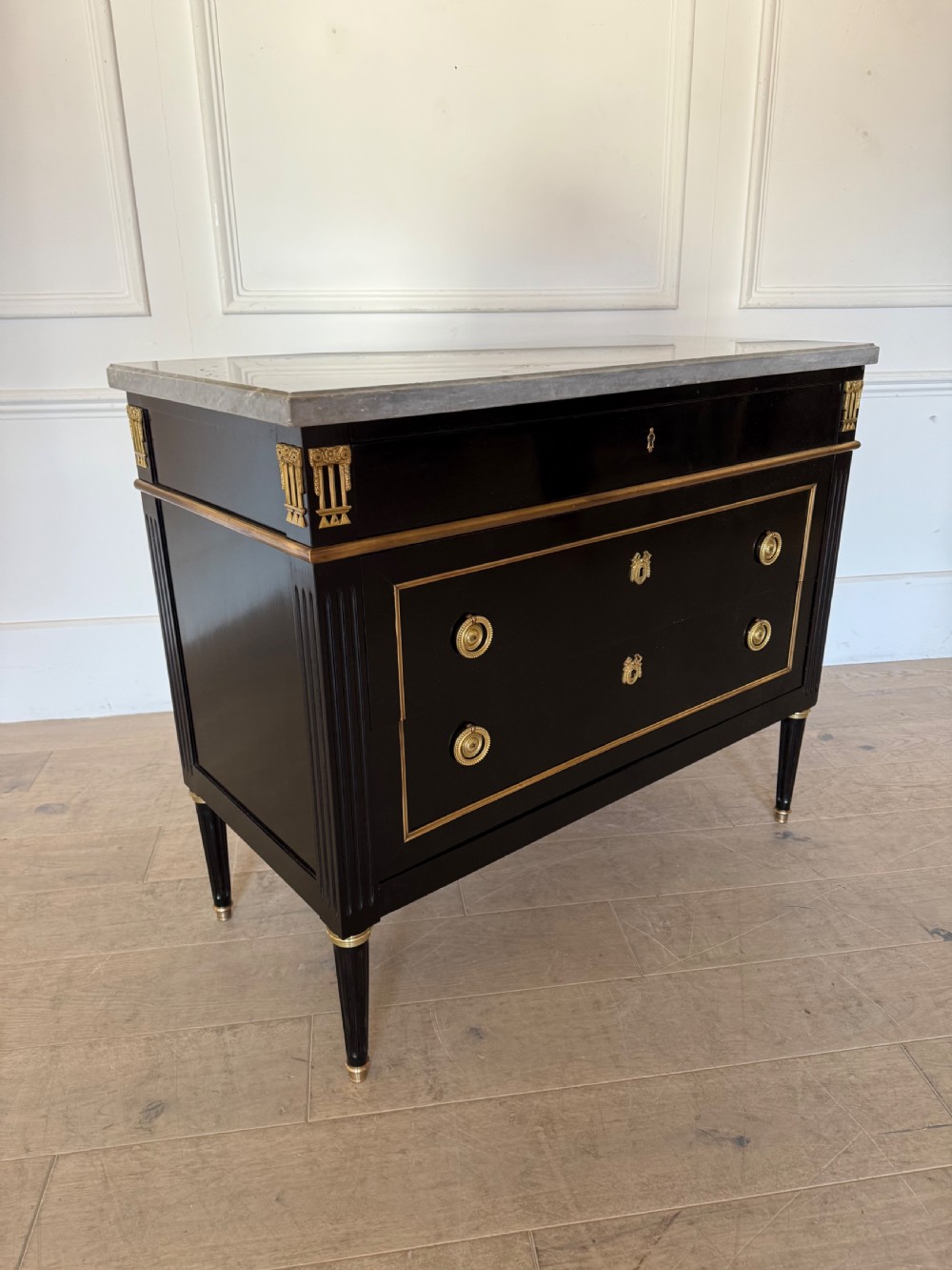 An ebonised commode chest of drawers featuring a grey marble top, brass ring handles, and gilt metal mounts. View VI