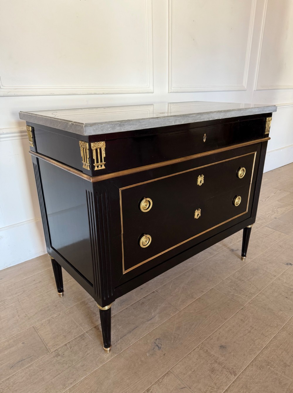 An ebonised commode chest of drawers featuring a grey marble top, brass ring handles, and gilt metal mounts. View VII