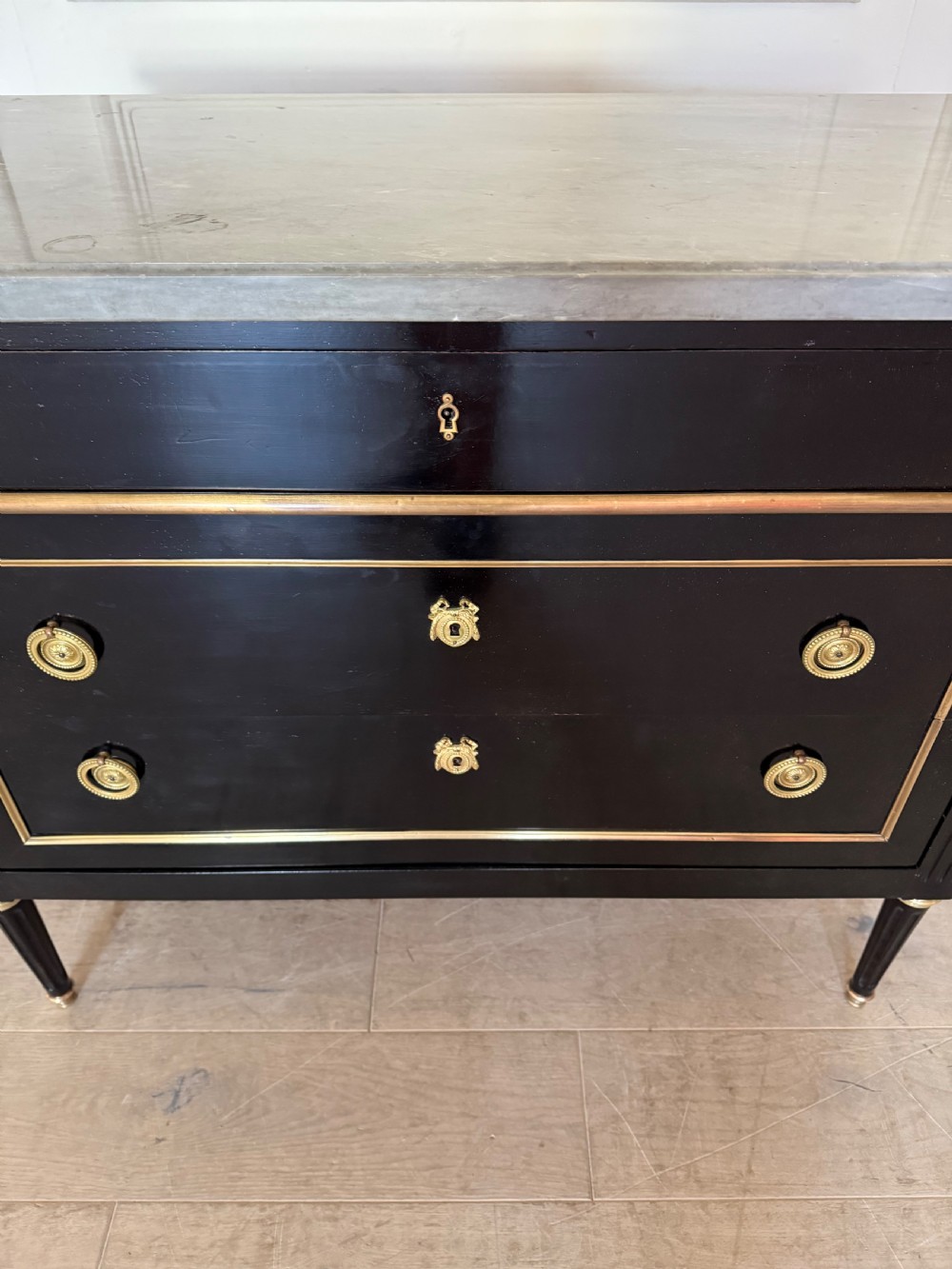 An ebonised commode chest of drawers featuring a grey marble top, brass ring handles, and gilt metal mounts. View VIII