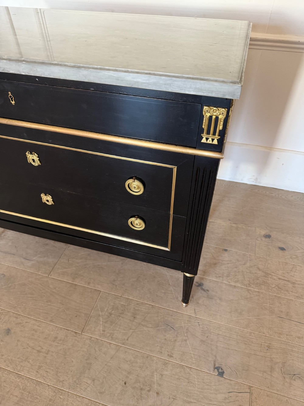 An ebonised commode chest of drawers featuring a grey marble top, brass ring handles, and gilt metal mounts. View IX