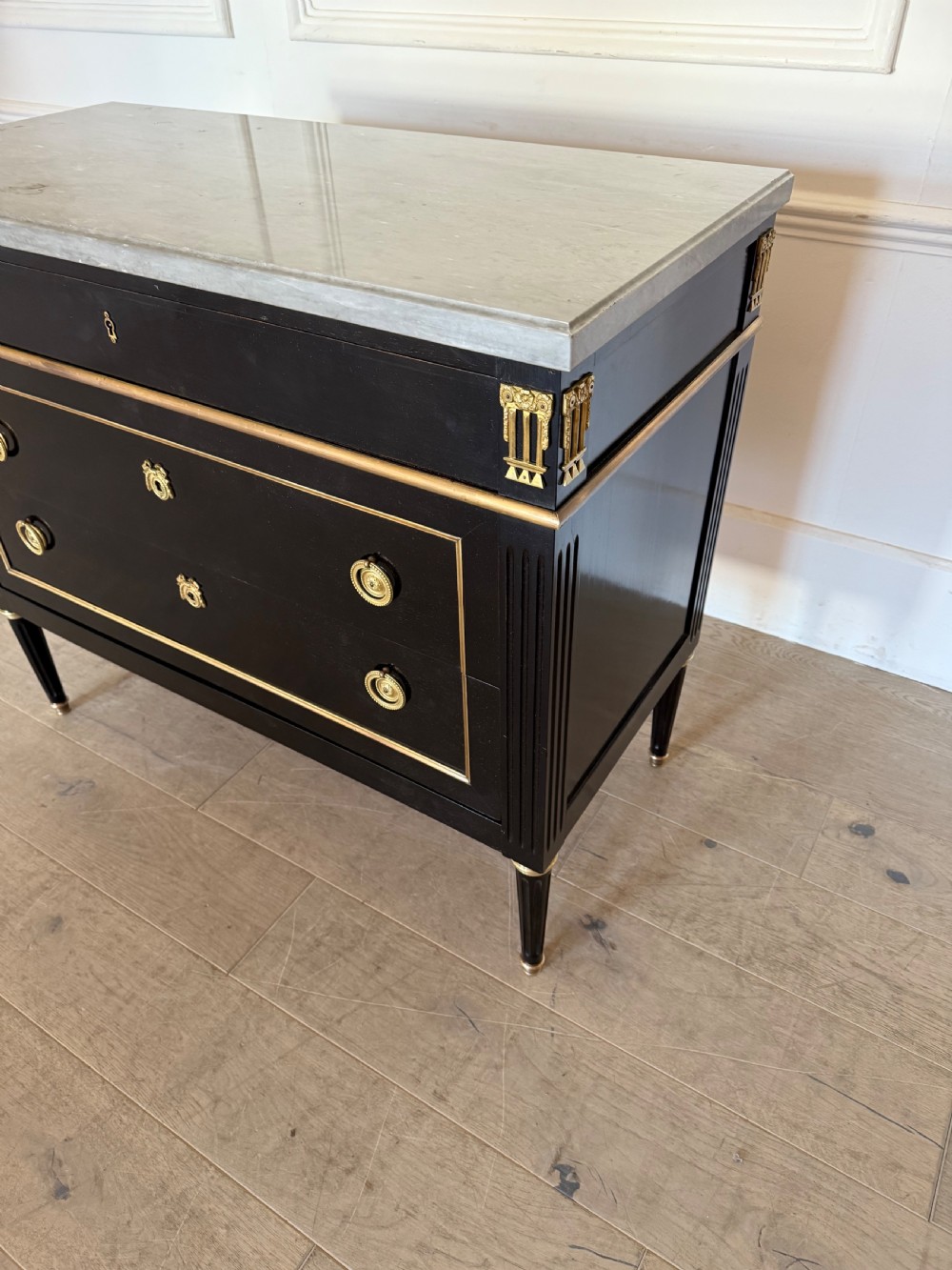 An ebonised commode chest of drawers featuring a grey marble top, brass ring handles, and gilt metal mounts. View X