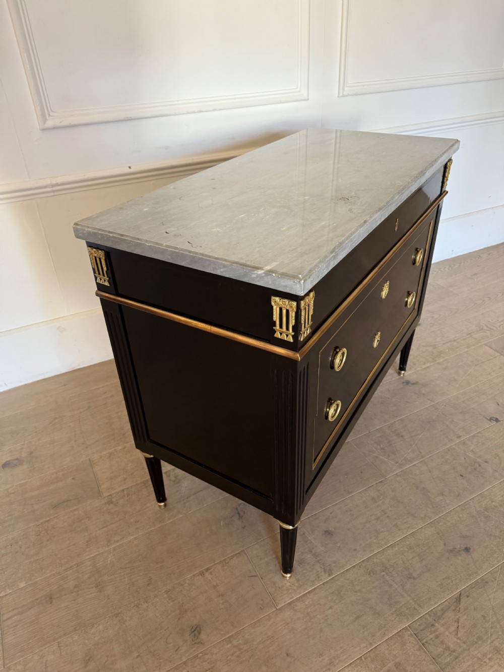 An ebonised commode chest of drawers featuring a grey marble top, brass ring handles, and gilt metal mounts. View XI