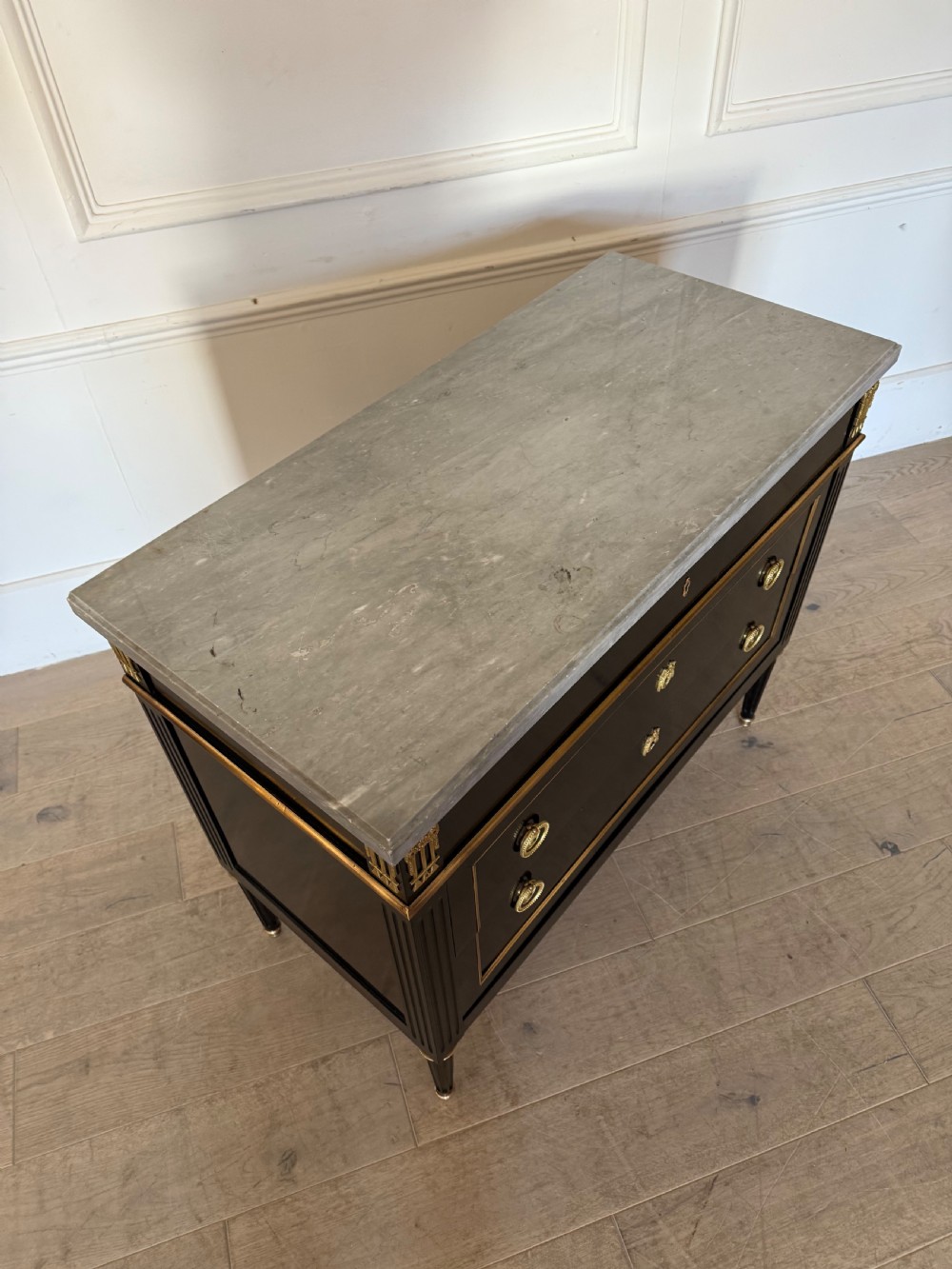An ebonised commode chest of drawers featuring a grey marble top, brass ring handles, and gilt metal mounts. View XII