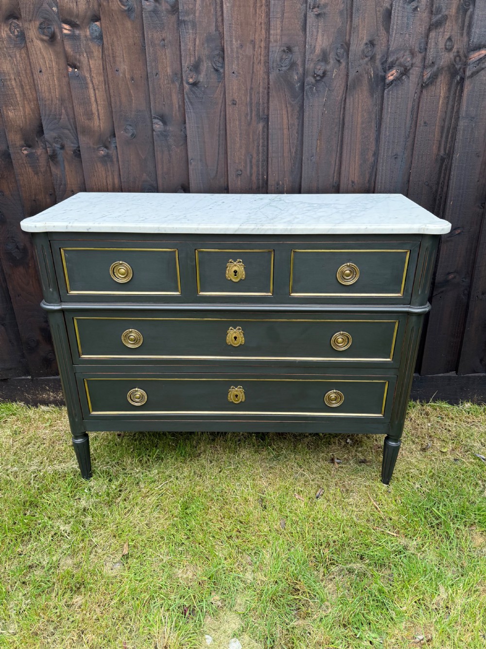 An early 20th-century painted Louis XVI style commode with a white marble top, dark green painted finish, gilded trim details, and cast brass ring pulls and keyhole escutcheons.