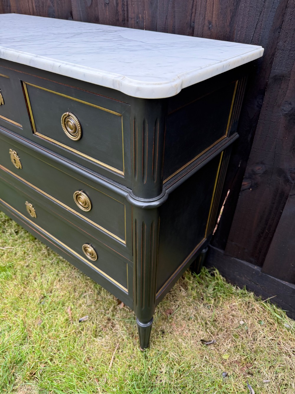 An early 20th-century painted Louis XVI style commode with a white marble top, dark green painted finish, gilded trim details, and cast brass ring pulls and keyhole escutcheons. View II