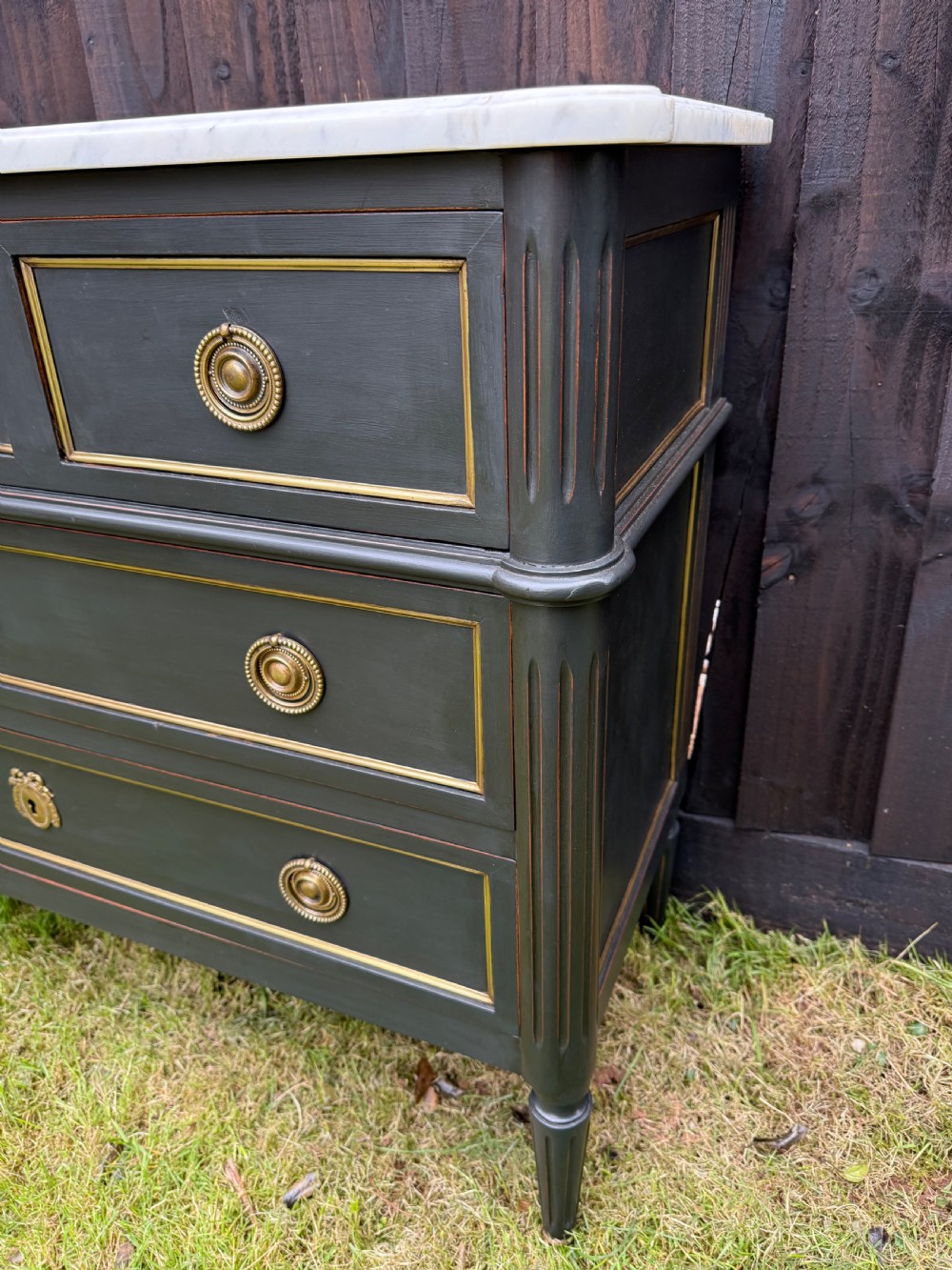 An early 20th-century painted Louis XVI style commode with a white marble top, dark green painted finish, gilded trim details, and cast brass ring pulls and keyhole escutcheons. View III